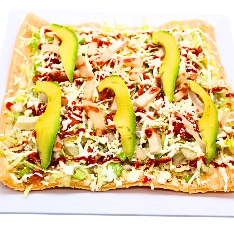 Crispy rectangular tostada loaded with lettuce, cheese, chicken, avocado, and drizzled sauces.