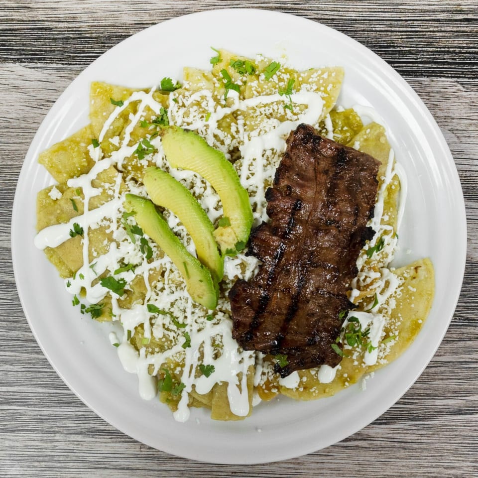 Chilaquiles verdes with grilled steak, avocado, crema, and crumbled cheese on a white plate.