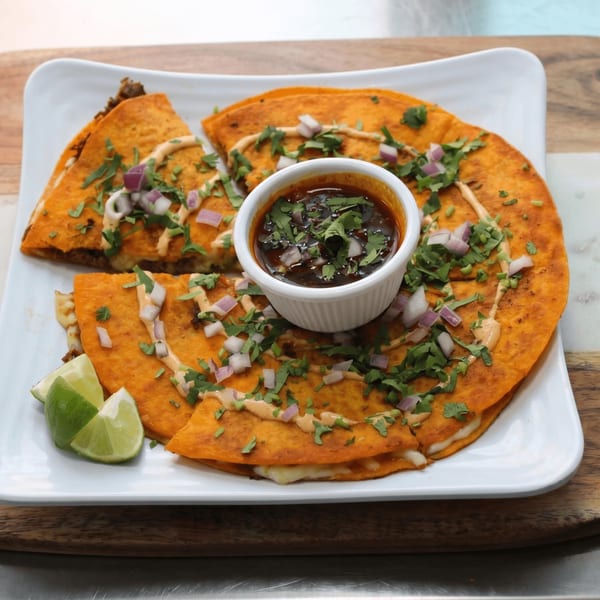 Birria quesadillas with cilantro, red onions, a side of consommé, and lime wedges on a white plate.