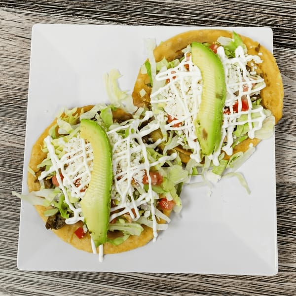 Two golden sopes topped with lettuce, cheese, tomatoes, sour cream, and avocado slices on a white plate.