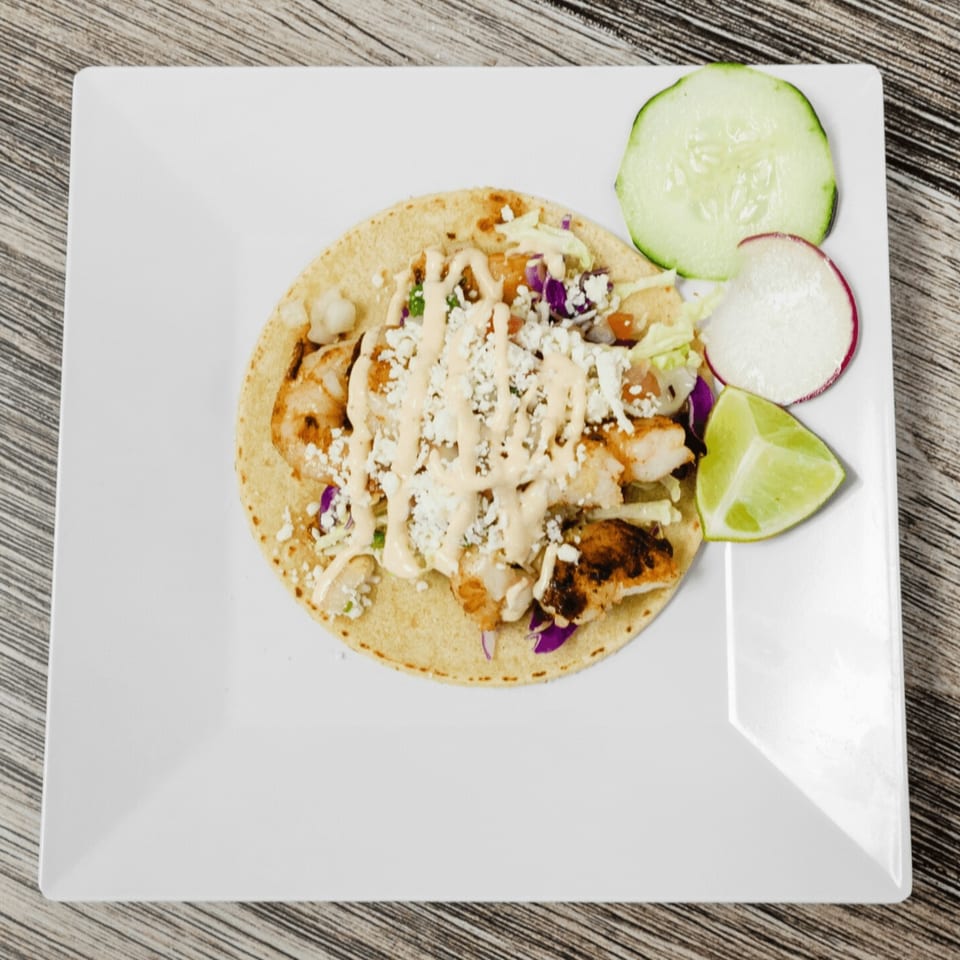 A shrimp taco with sauce, cheese, and cabbage, served with cucumber, radish, and lime.
