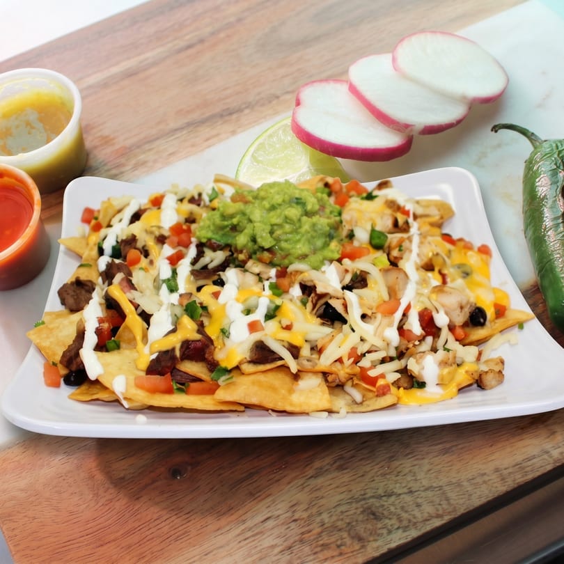 Loaded nachos with meat, cheese, guacamole, and sour cream, served with sauces and garnishes.