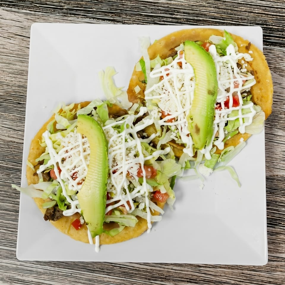 Two golden sopes topped with lettuce, cheese, tomatoes, avocado, and a drizzle of cream.