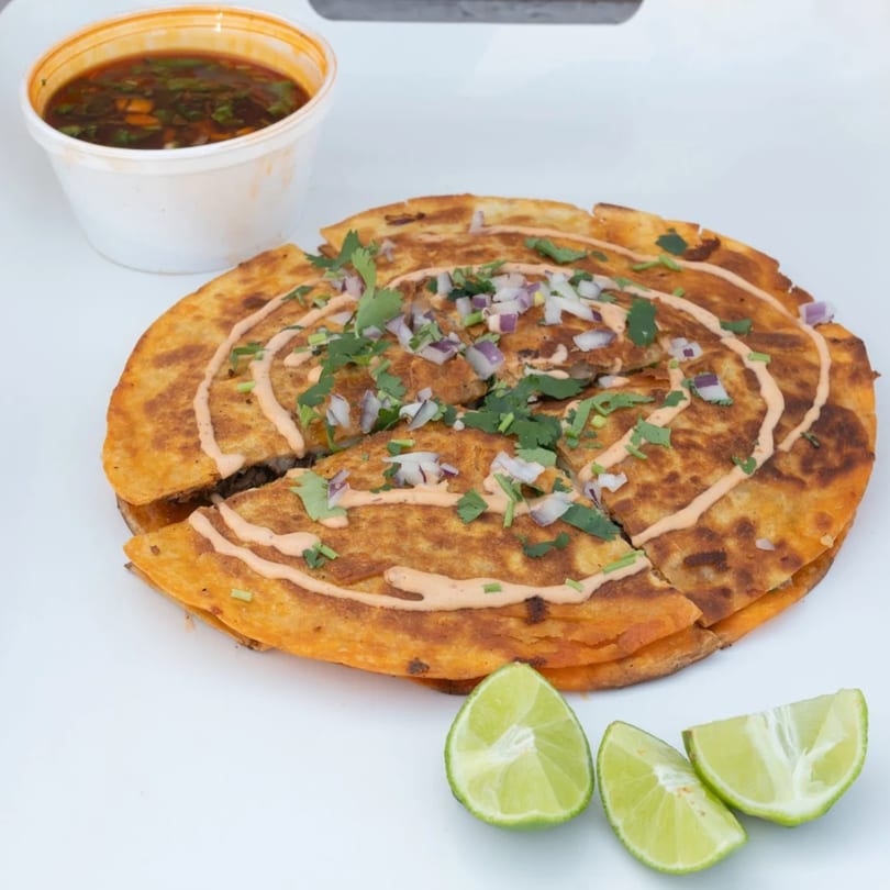 Birria quesadilla with red onions, cilantro, and sauce, served with consome and lime wedges.