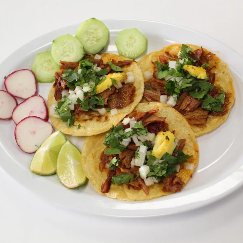 Three al pastor tacos with pineapple, cilantro, and onions, served with radishes, cucumbers, and lime.
