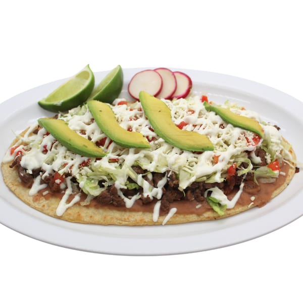 A large Mexican sope topped with meat, beans, lettuce, cheese, avocado, and sour cream, garnished with lime and radish.