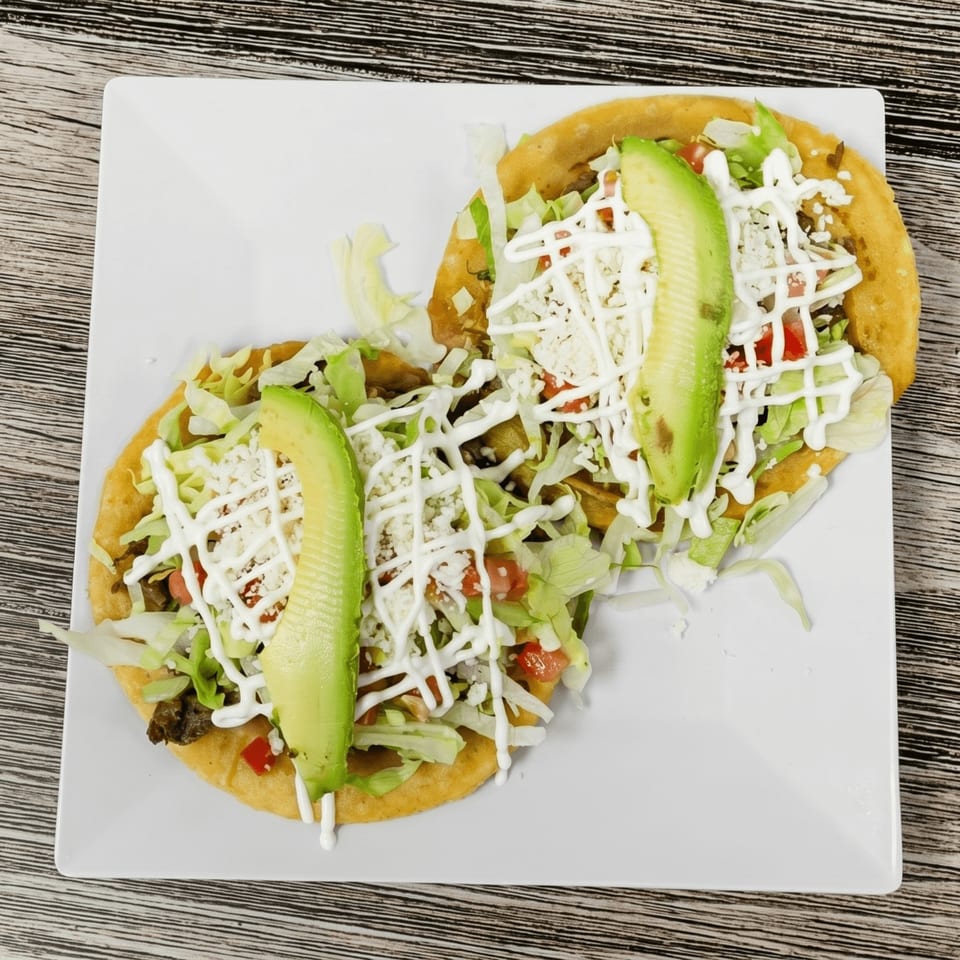 Two golden sopes topped with lettuce, cheese, tomatoes, sour cream, and avocado slices on a white plate.